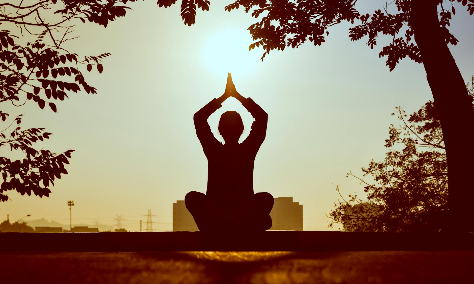 Someone meditates with their hands together over their head with a setting sun in the background.
