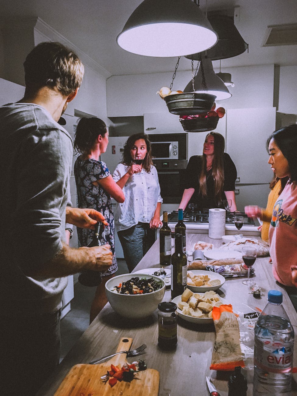 A group of four people socialize around a kitchen table filled with food and drinks.
