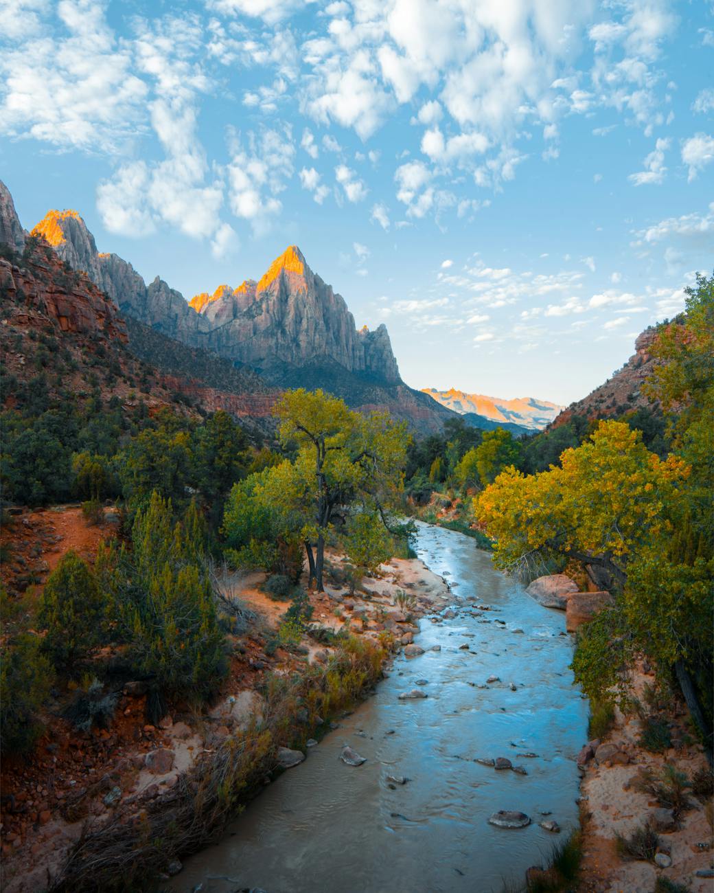 A river lined with trees and mountaintops in the background against a blue sky filled with clouds.