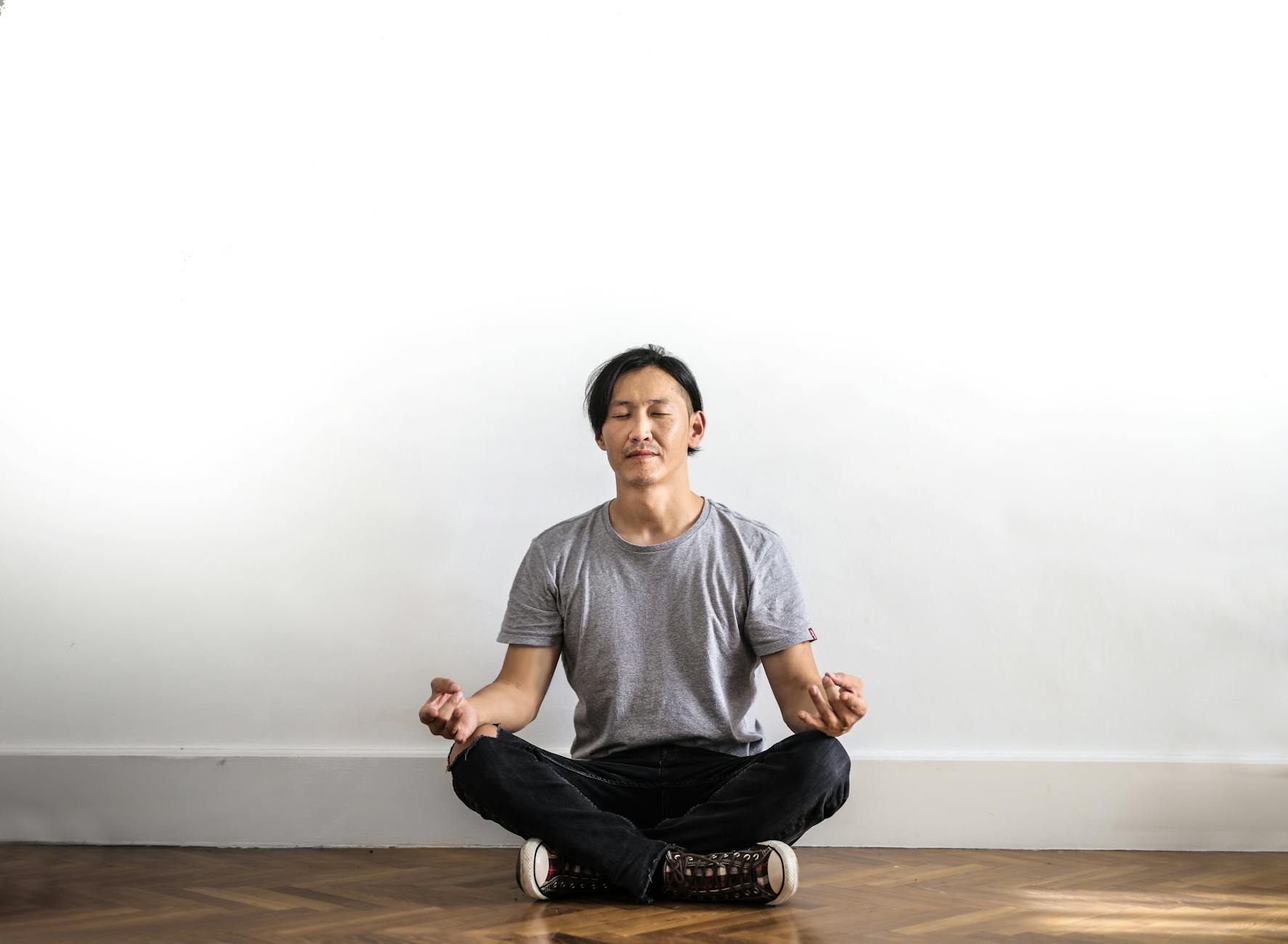 A person meditates on a wooden floor against a white wall.