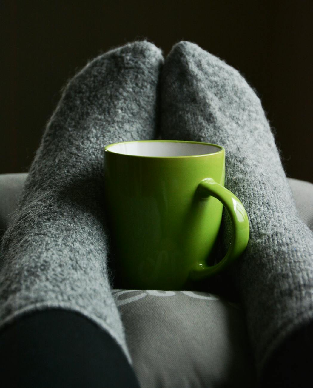 A pair of feet in grey socks hold a green mug on top of a grey pillow.
